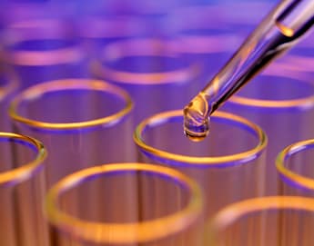 A close-up image shows a pipette dispensing a clear liquid into one of several glass test tubes. The background is blurred with a mix of blue and purple hues, creating a scientific and vibrant atmosphere.