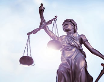 A bronze statue of Lady Justice holding scales in her left hand and a sword in her right hand, with a blindfold covering her eyes. The background is a clear, bright blue sky with light clouds. The image focuses on the statue, symbolizing fairness and law.