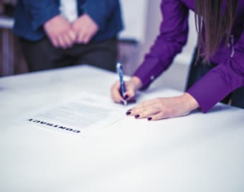 A person wearing a purple shirt is signing a contract on a white table with a pen. Another person in a blue suit stands opposite, with only their torso and hands visible. A small white coffee cup is placed on the table in the background.