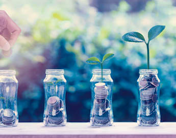 A hand places a coin into one of four glass jars filled with coins. Two jars have small plants sprouting from them, symbolizing financial growth and investment. The background is blurred greenery, highlighting the focus on the jars and coins.