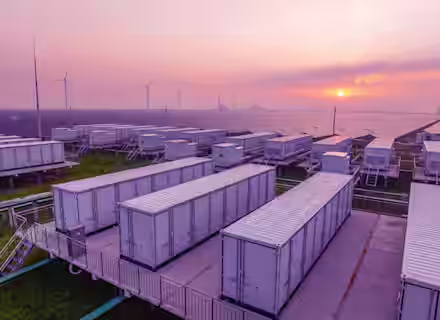 Large white battery storage units are arranged in rows at a renewable energy facility, with solar panels and wind turbines in the background under a purple and pink sunset sky.