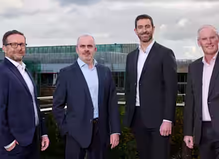Four men in business attire stand together on a balcony outdoors, with a modern glass building and cloudy sky in the background. They are smiling and facing the camera.