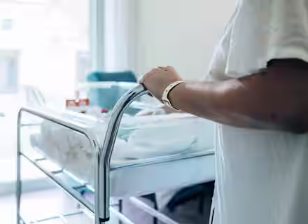 A person in a hospital gown with an ID bracelet stands next to a hospital bassinet with a baby inside, covered in a white blanket. The room is softly lit, and a window is visible in the background.