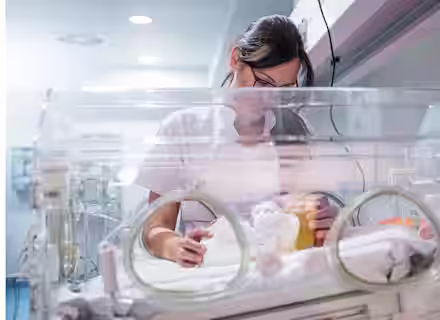 A healthcare professional tending to a baby in an incubator in a neonatal unit. The adult is wearing glasses and a white uniform, and the scene suggests a hospital environment with medical equipment visible.