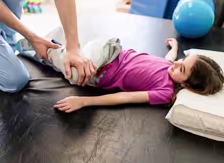 A physical therapist helps a young girl lying on a table by gently stretching her leg. The girl is wearing a pink shirt and gray pants, and she rests her head on a pillow. Exercise equipment is seen in the background.