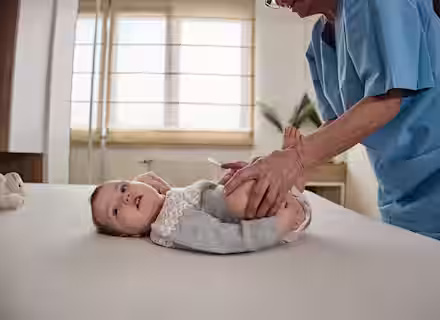 A baby lies on an examination table while an adult in blue scrubs gently holds and moves the baby’s legs, likely during a medical or physical therapy checkup in a bright room.