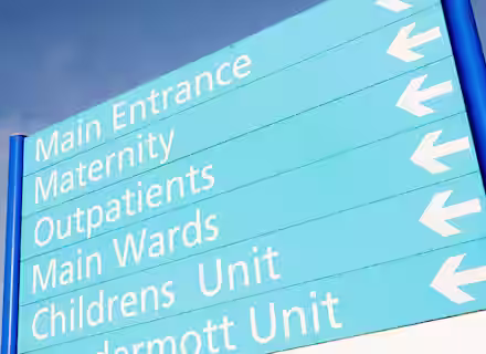 A blue hospital directional sign with white text and arrows pointing left, listing Main Entrance, Maternity, Outpatients, Main Wards, Childrens Unit, and part of another unit name.