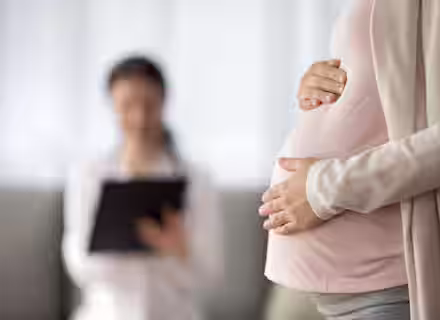 A pregnant woman gently holds her belly while standing in the foreground. In the blurred background, a healthcare professional sits and writes on a clipboard.