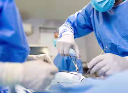 Surgeons, dressed in blue surgical gowns and gloves, perform an operation. One surgeon holds forceps, while another assists. The sterile environment and focused attention on the task are evident, highlighting the precision and care involved in the procedure.