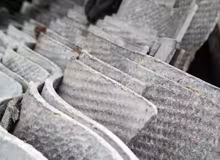 Stacked corrugated sheets of weathered, gray asbestos roofing material, showing a rough texture with visible fibers. The sheets are densely packed, illustrating an industrial or construction setting.