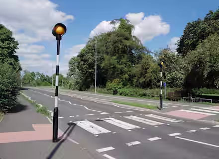 A pedestrian zebra crossing with black and white poles topped by amber lights on a quiet road, surrounded by green trees and bushes under a partly cloudy blue sky.