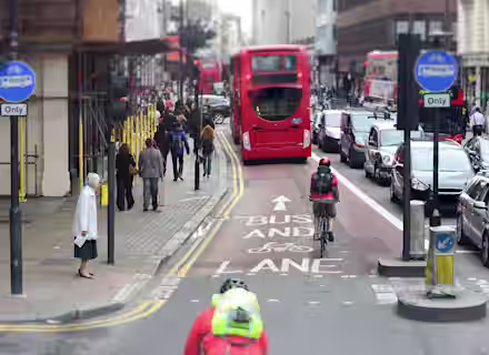 A busy city street with a dedicated bus and bicycle lane. A red double-decker bus is moving down the lane, followed by a bicyclist wearing a helmet and backpack. Pedestrians are walking on the sidewalk, and various vehicles are on the adjacent car lane.