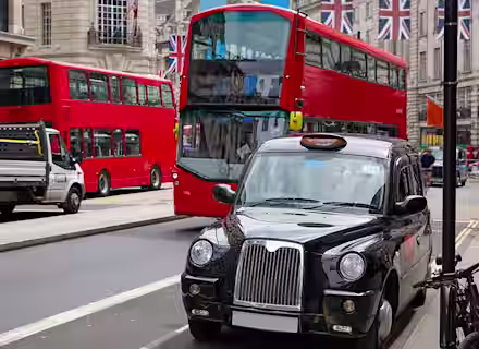 A busy urban street scene featuring two red double-decker buses, a traditional black taxi, and a service truck. The background showcases buildings adorned with British flags, indicating a vibrant city atmosphere.