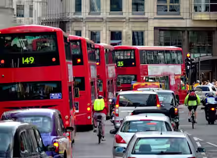 A busy city street with red double-decker buses, cars, and cyclists. Several buses are lined up in traffic, each displaying different route numbers. Buildings line the street background, and people are visible on the sidewalks.