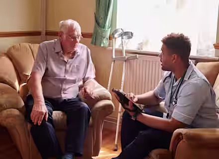 An elderly man sitting on a sofa talks with a healthcare professional kneeling beside him. The professional holds a tablet, wearing a stethoscope, with a crutch seen in the background. They are in a warmly lit living room.