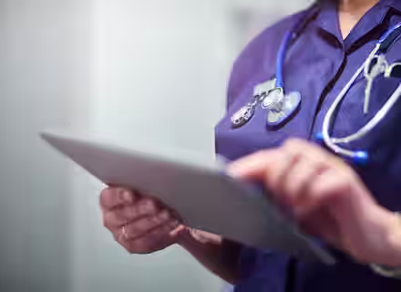 A healthcare professional in a navy blue uniform holding and using a digital tablet. A stethoscope is draped around their neck, and the background is softly blurred.