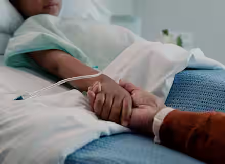 A child lies in a hospital bed with an IV in their arm, holding hands with an adult who sits beside them, offering comfort and support.