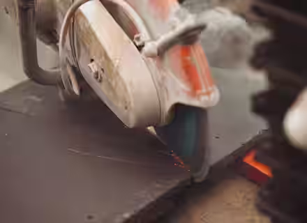 A close-up of a circular saw cutting through a dark sheet of material, with sparks flying from the blade. The scene appears to be part of a construction or fabrication process.