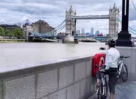 A cyclist in casual wear leans on a stone railing, gazing toward the Tower Bridge in London. The cyclist's bike is resting nearby, and the cloudy sky forms the backdrop over the bridge and river.
