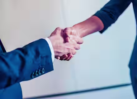 Close-up of two people shaking hands. One person is wearing a dark blue suit with a white shirt, while the other person is wearing a dark blue jacket. The background is blurred and light-colored, suggesting a professional or business setting.