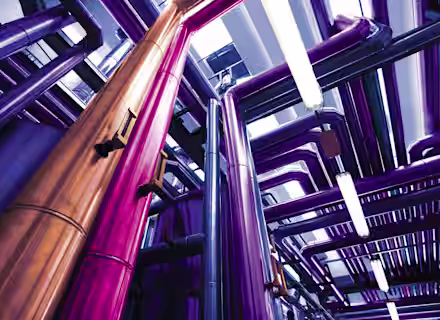 A vibrant and colorful industrial scene with tall metallic pipes in shades of purple, pink, and copper running in various directions under bright lighting. The pipes are part of a complex system, creating a visually striking and futuristic environment.