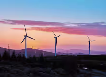 A scenic view of multiple wind turbines scattered in a hilly landscape during sunset. The sky is painted in vibrant hues of pink, orange, and purple, with silhouettes of distant mountains and a foreground dotted with trees and rocks.