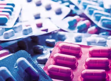 Several packs of pills in various shapes and colors are scattered on a surface. The medication is contained in blister packs, with some capsules being half white and half blue, others in different color combinations. The image uses blue and pink lighting.