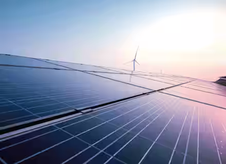 Close-up of solar panels with a wind turbine in the background under a clear sky. The sun is shining brightly, reflecting off the panels, representing renewable energy sources.