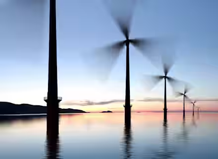 A row of wind turbines standing in calm water during sunset, with blurred blades indicating movement. The sky transitions from light blue to peach and pink near the horizon. Hills are faintly visible in the background.
