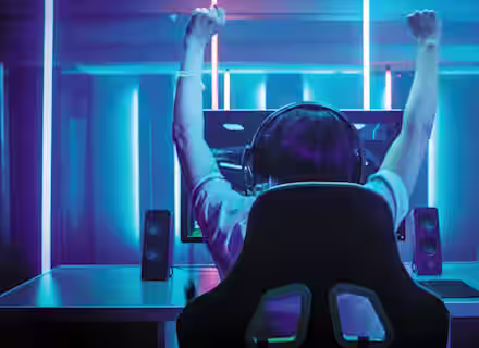 A person with headphones, viewed from behind, raises their arms in victory while sitting at a desk in front of a computer monitor. The room is dimly lit with blue and pink neon lights, creating a cyber atmosphere.