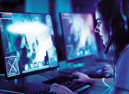 A person wearing a headset sits at a computer, engaged in a video game. The room is dimly lit with a blue hue. Multiple monitors display gameplay, featuring vibrant graphics. The person uses a keyboard and mouse, focused on the screen.