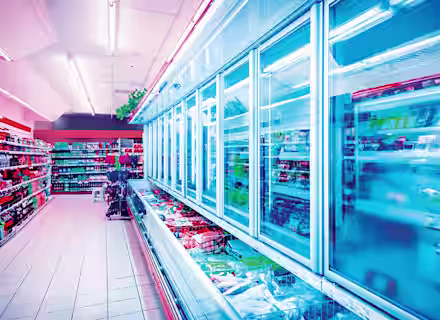 A brightly lit supermarket aisle showcases refrigerated freezer sections with glass doors. Shelves are stocked with various frozen items, and adjacent shelves display other grocery products. The floor is clean, tiled, and the store appears well-organized.