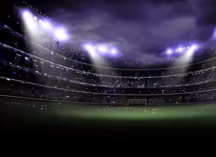 A dimly lit, large football stadium at night with bright floodlights illuminating the field. The stands are filled with spectators, and the atmosphere is vibrant. Confetti can be seen falling, creating a celebratory scene, while the green pitch is ready for action.