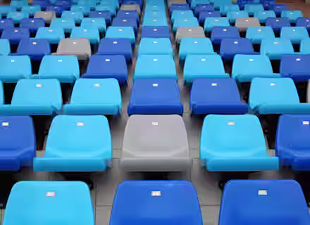 An empty stadium seating area with rows of plastic seats. The seats are predominantly blue, with a few gray ones interspersed randomly. Small white tags are visible on the backs of each seat.