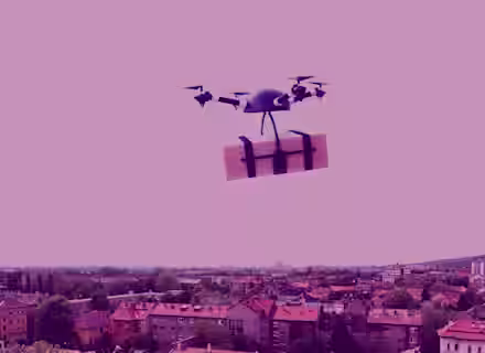 A drone is flying over a cityscape, carrying a package secured with black straps. The image has a pink overlay that tints the entire scene, including the buildings below and the sky above.