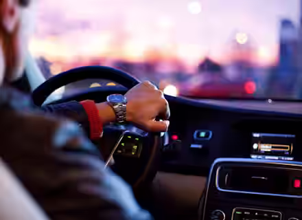 A person is driving a car, visible from behind the passenger seat. The driver wears a watch on their left wrist. The dashboard displays various controls and a digital screen. Outside the car, the sky is colorful with pinks and purples, indicating dawn or dusk.
