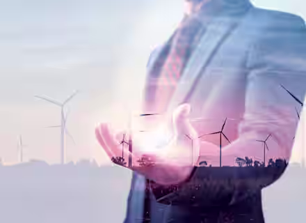 A double exposure image showing a person in a suit with an extended hand, overlaid with wind turbines in the background. The person appears to be holding a bright light, symbolizing renewable energy and sustainability.