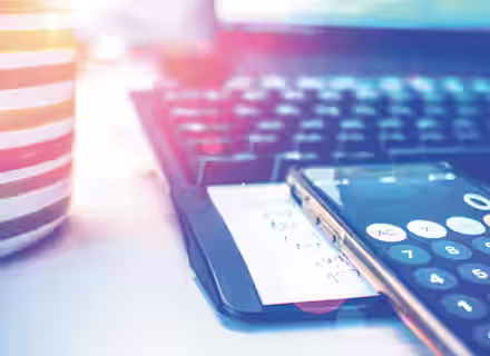 A close-up of a desk workspace with a striped mug, a computer keyboard, and a smartphone displaying a calculator app. A piece of paper with handwritten notes is beneath the phone. Sunlight creates lens flare effects on the image.