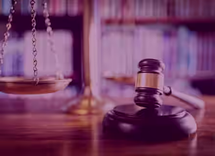 A wooden gavel rests on a sound block in the foreground, with a balanced scale of justice in the background. The setting appears to be a courtroom or a law library, indicated by the blurred shelves filled with books.