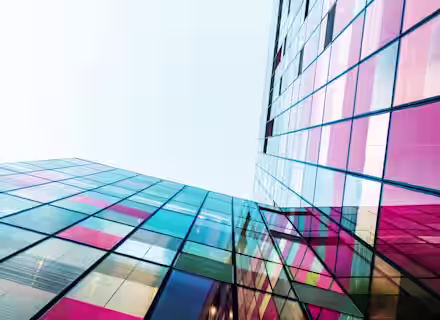 A low-angle view of modern glass buildings reflecting colorful hues of pink, red, blue, and yellow under a clear sky. The structures create a geometric pattern with their intersecting lines and vibrant panels.