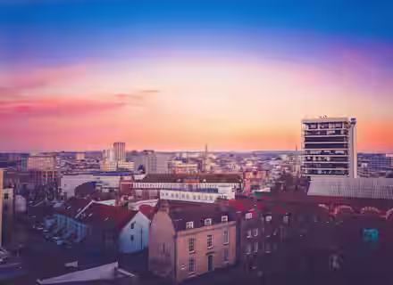 A cityscape at dusk with a vibrant sky transitioning from deep blue at the top to soft orange near the horizon. Various buildings, including modern high-rises and older structures with red-tiled roofs, are visible. The city is bathed in a warm, soft light.