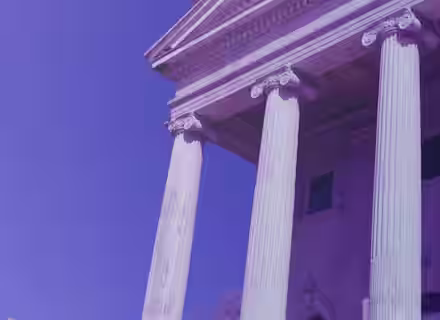 A classic building with tall Ionic columns and a triangular pediment is shown from a low angle against a clear blue sky, highlighting the architectural details. The photograph has a purple tint cast over it.