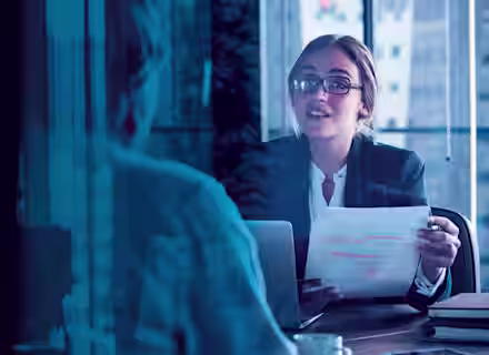 A woman wearing glasses and a business suit is holding a paper and speaking to another person across a desk in an office setting. The scene is viewed through a window, reflecting the cityscape outside.