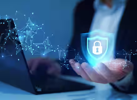 A person in a suit holds a digital shield with a padlock symbol above their hand, representing cybersecurity, in front of a laptop with abstract blue network connections.