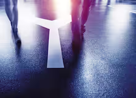 Two people are walking on a wet street at night, illuminated by streetlights. They are approaching a large painted Y-shaped arrow on the ground, which splits into two directions. The scene has a moody, reflective atmosphere.