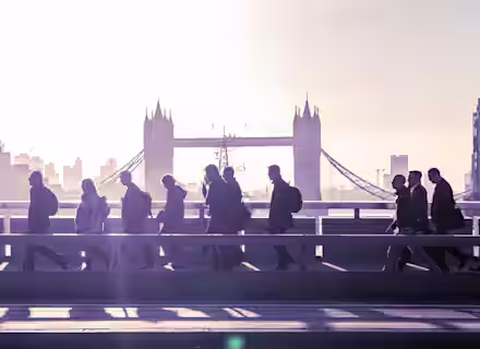Silhouettes of people walking across a bridge during early morning with the iconic Tower Bridge in London visible in the background. The sky is filled with a soft glow from the rising sun.
