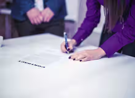 A person wearing a purple shirt is signing a contract on a white table with a pen. Another person in a blue suit stands opposite, with only their torso and hands visible. A small white coffee cup is placed on the table in the background.