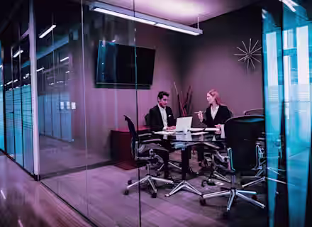 Two people in business attire sit at a conference table in a modern glass-walled office, working on a laptop and discussing documents. The room features a TV, modern decor, and office chairs.