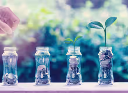 A hand places a coin into one of four glass jars filled with coins. Two jars have small plants sprouting from them, symbolizing financial growth and investment. The background is blurred greenery, highlighting the focus on the jars and coins.
