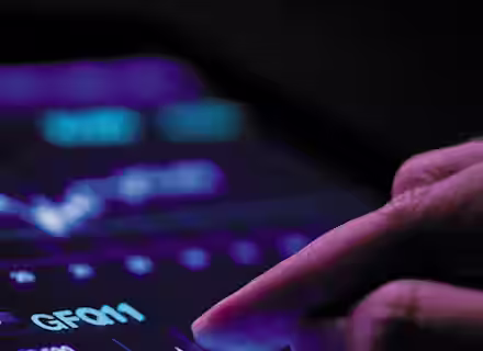 A person taps a tablet screen displaying a digital stock trading interface with various graphs, charts, and numbers lit up in neon purple and blue against a dark background. The prominent graph shows upward and downward trends.